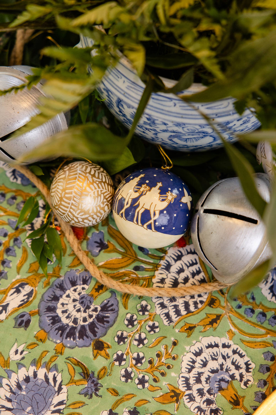 Decorative items including a blue ornament with gold deer, a silver bell, and a gold ornament on a patterned fabric background.